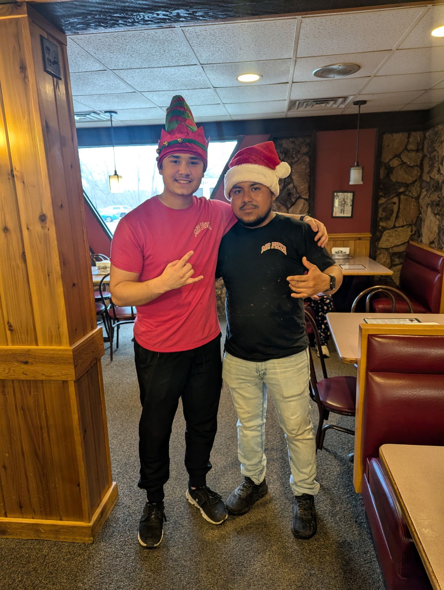 Two people in festive hats posing together in a restaurant booth area, smiling and holding drinks.