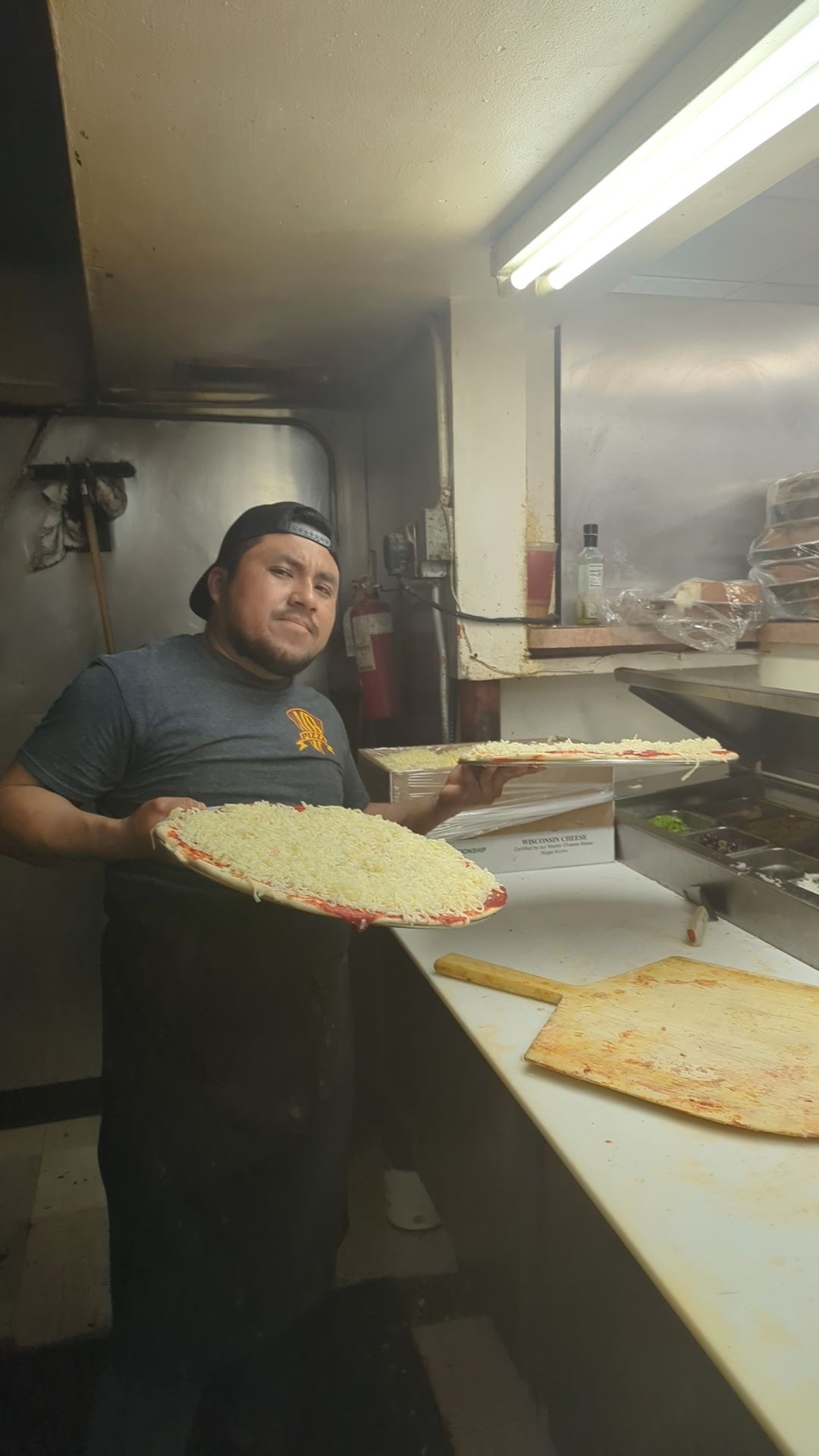 Person holding a large pizza in a commercial kitchen with pizzas on a prep counter