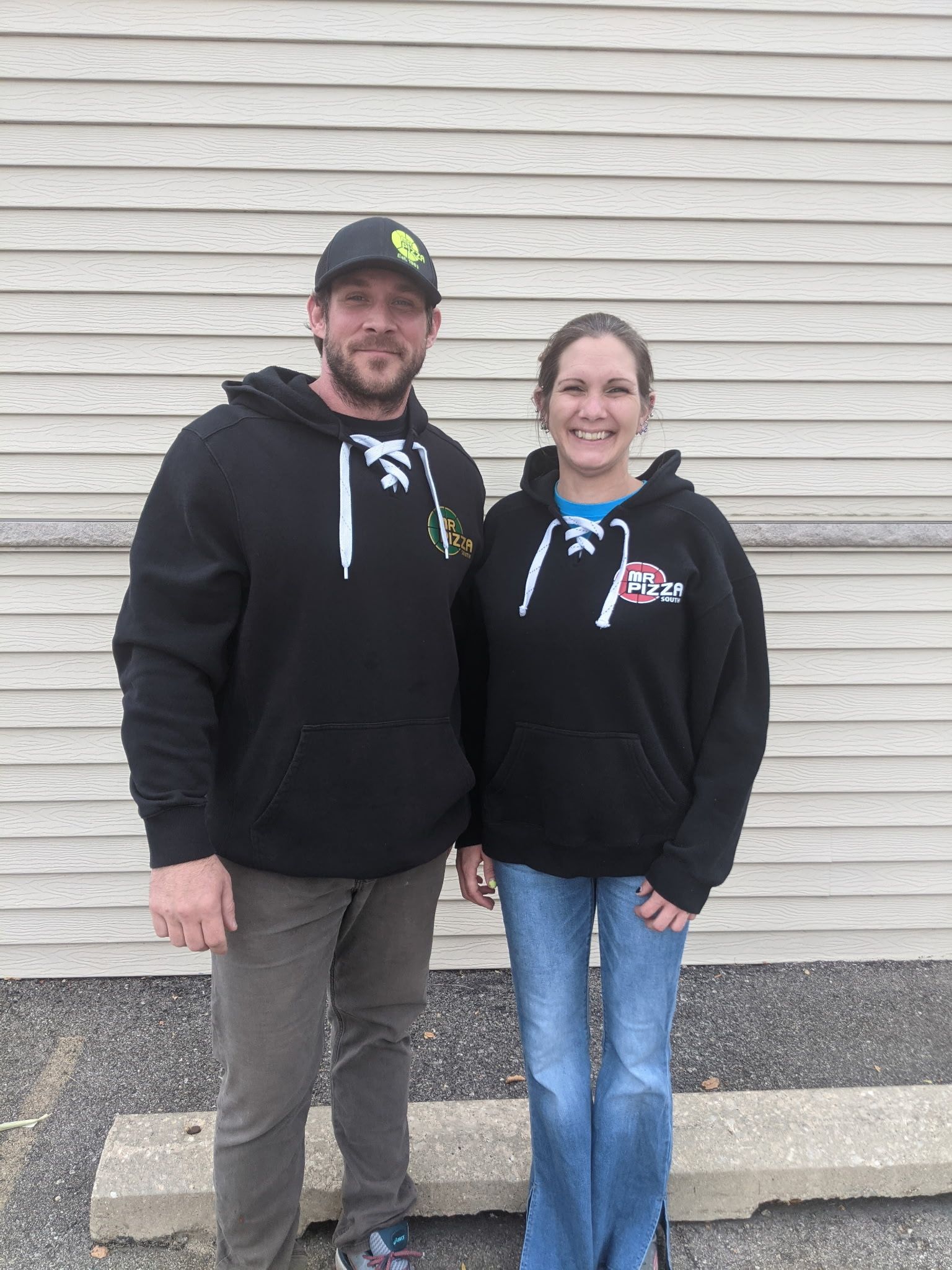 Two people in black hoodies standing side by side against a beige siding wall