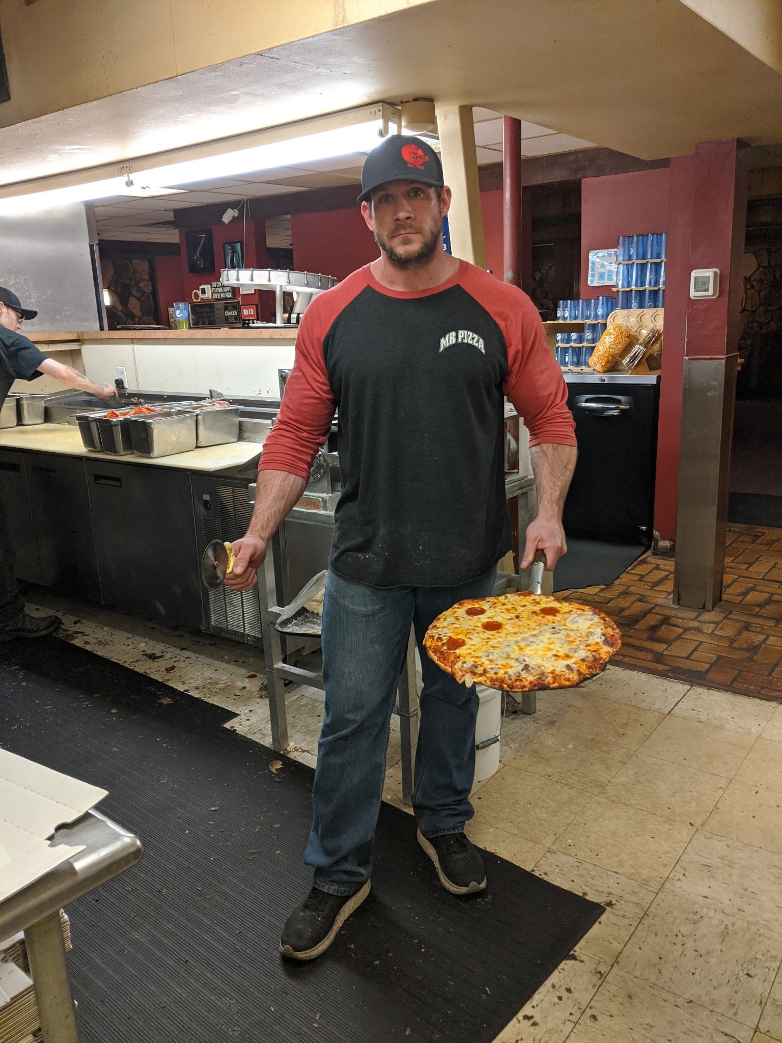 Man holding a pizza peel with a large pizza in a restaurant kitchen, wearing a black-and-red shirt and cap