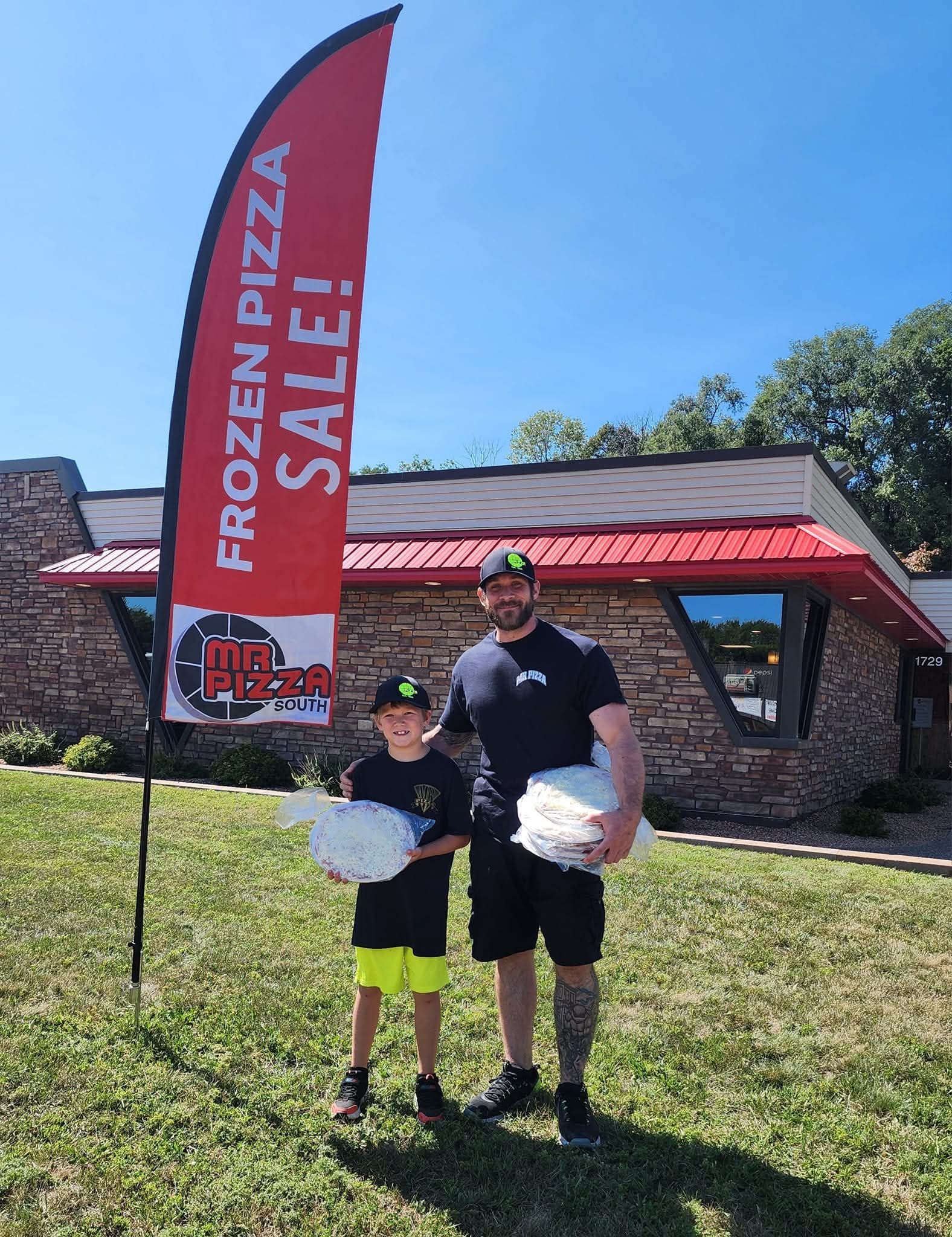 Two people holding frozen pizzas beside a red sale flag and building under a blue sky.