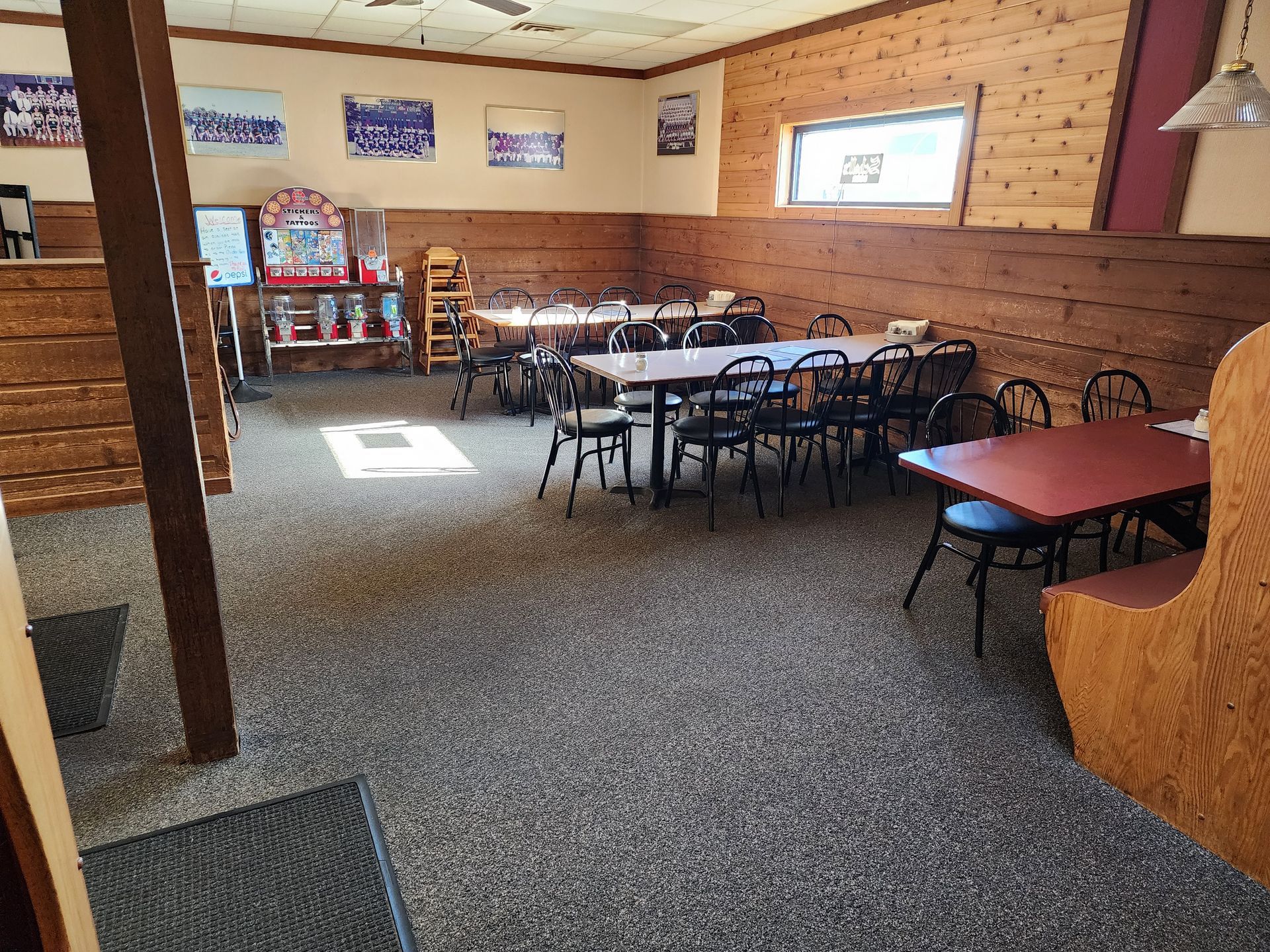 Empty restaurant dining room with scattered tables and chairs, wood-paneled walls, and sunlight on carpeted floor