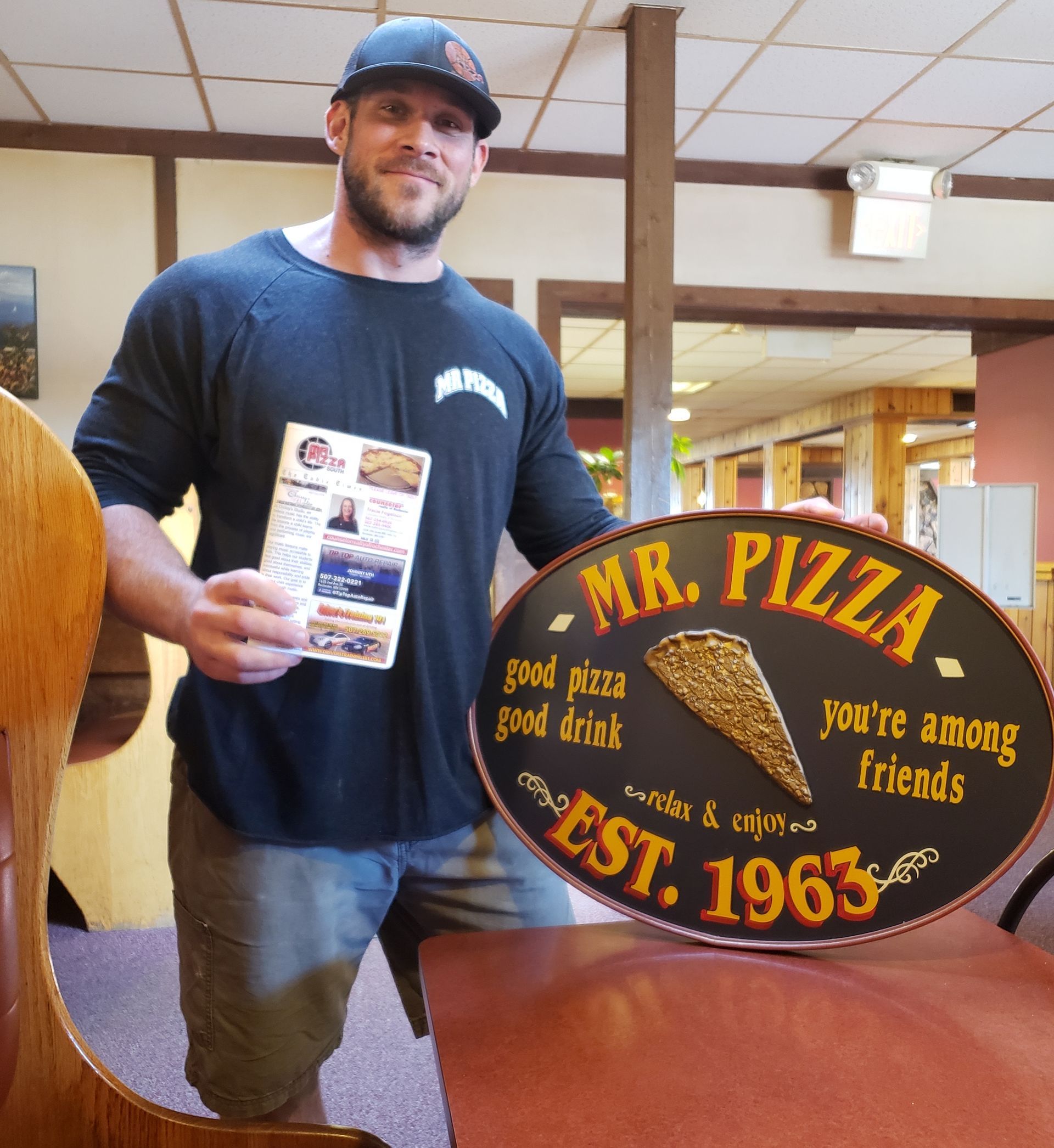 Man holding a Mr. Pizza sign and flyer inside a restaurant, with a large pizza sign nearby.