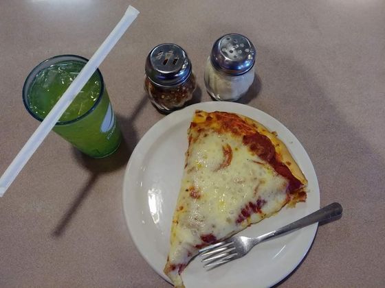Slice of cheese pizza on a white plate with a fork, beside a green drink and two condiment shakers.
