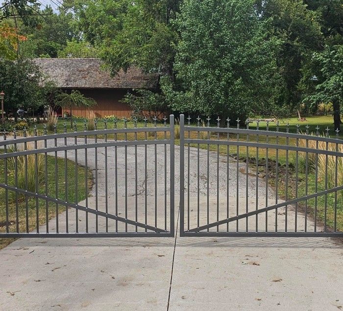 Gray metal gate, arched, set on a concrete driveway, leading to a house with a brown roof and surrounded by trees.