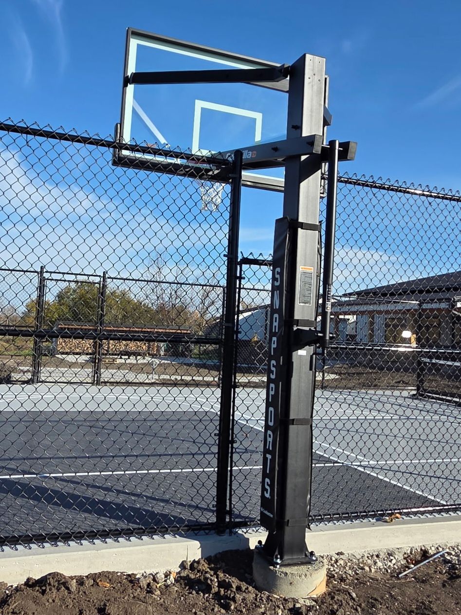 Basketball hoop on a black post at an outdoor court, behind a chain link fence.