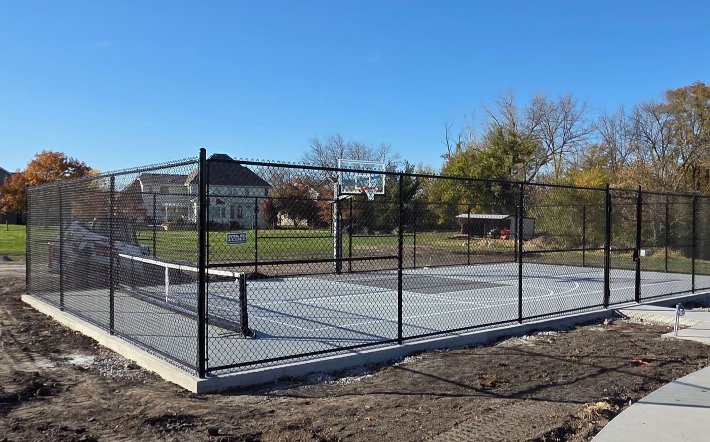 Outdoor basketball court enclosed by a black chain-link fence on a sunny day.