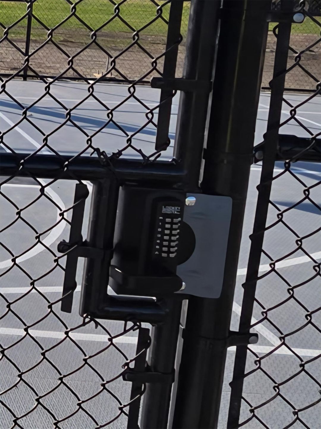 Black gate with a keypad lock on a chain link fence, with a basketball court in the background.