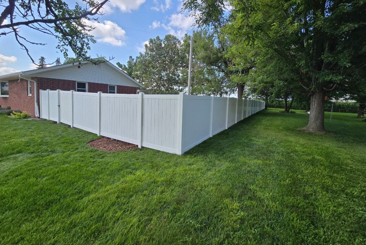 White vinyl fence surrounding a grassy yard, next to a brick house and trees under a blue sky.