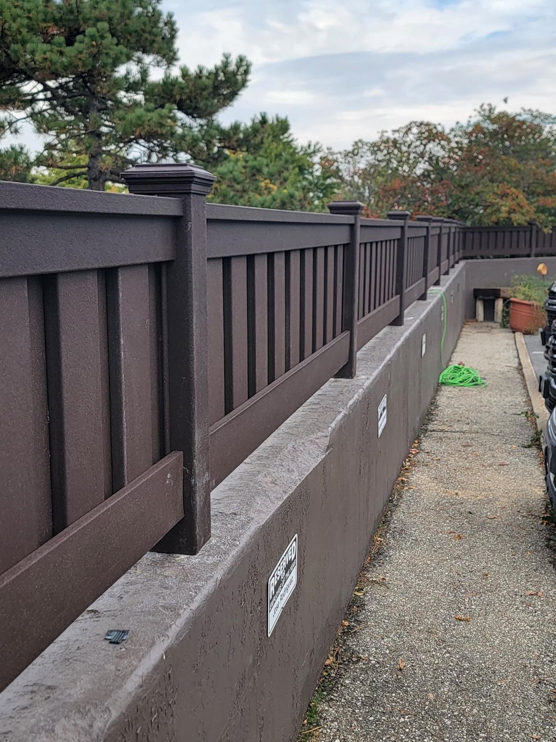 Brown fence atop a low concrete wall, gravel path alongside, with greenery in the background.