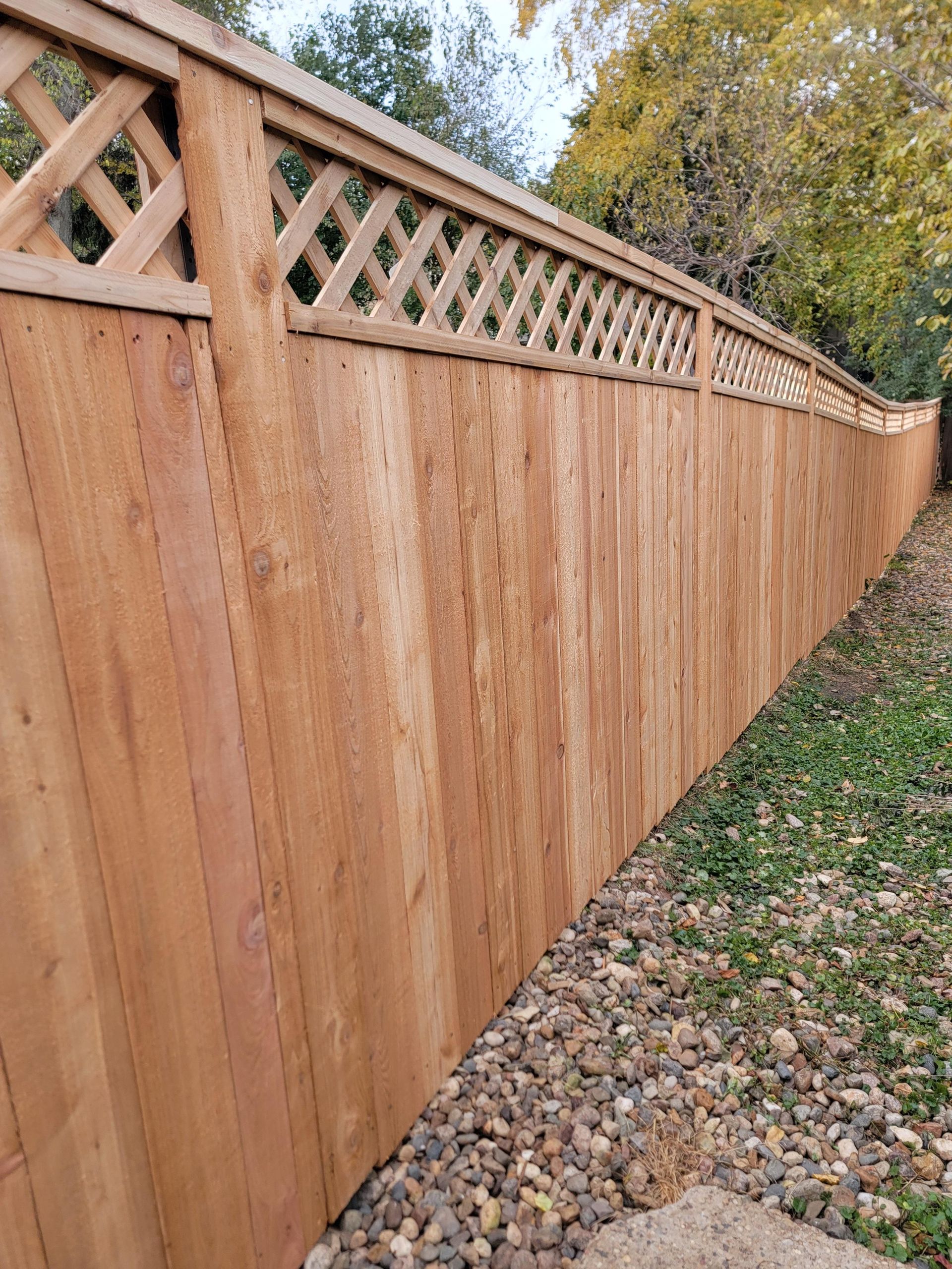 Wooden fence with lattice top in a yard with small rocks and green grass.