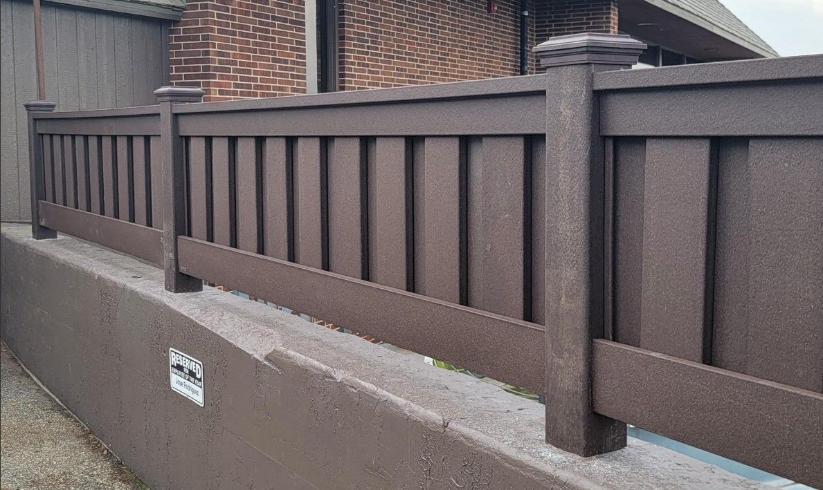 Brown composite fence on a concrete wall; brick building in background.