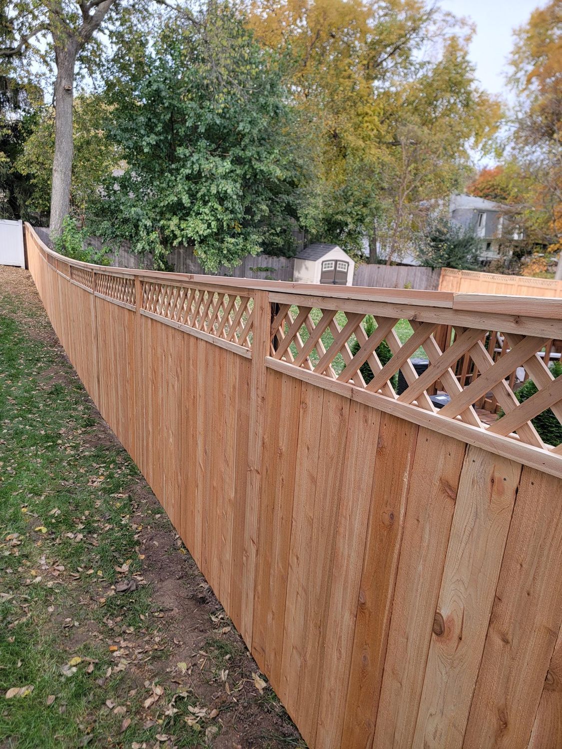 Wooden fence with lattice top running along a green lawn, trees, and houses in the background.
