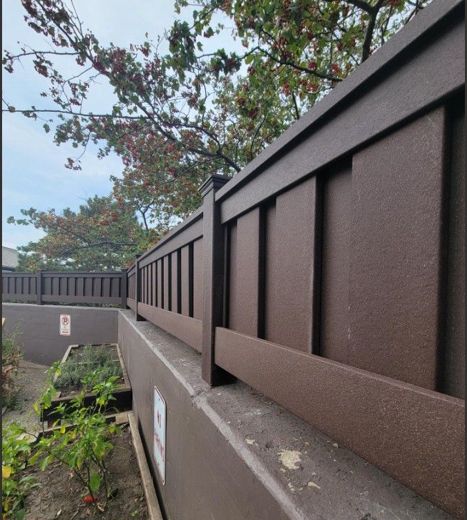 Brown fence atop a retaining wall, with plants and trees visible in the background.