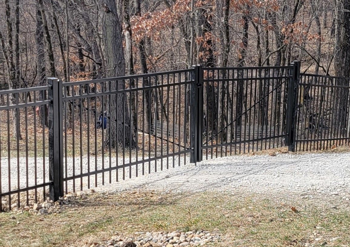 A black metal fence surrounds a gravel road in the woods.