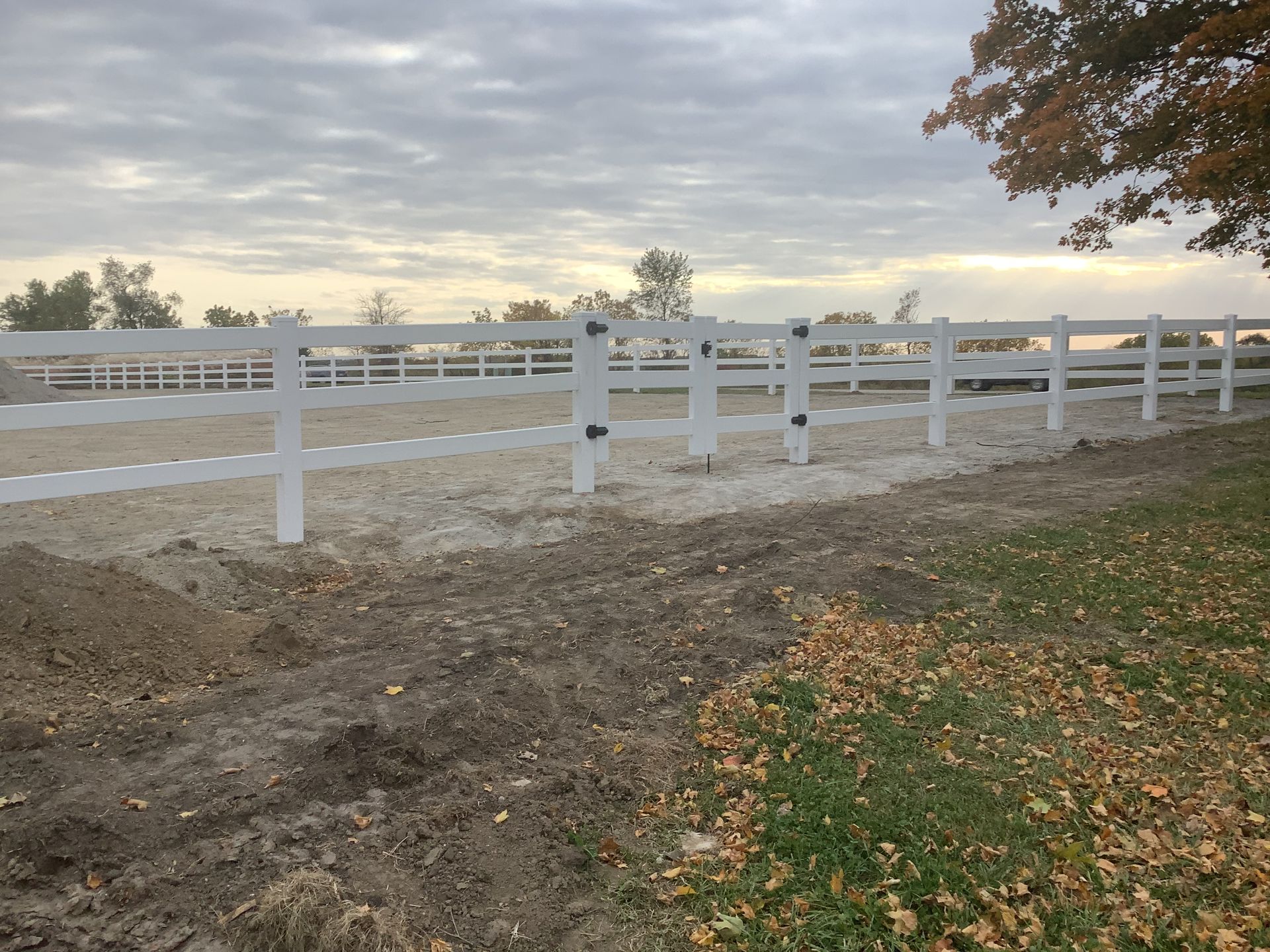 A white fence surrounds a dirt field with a tree in the background