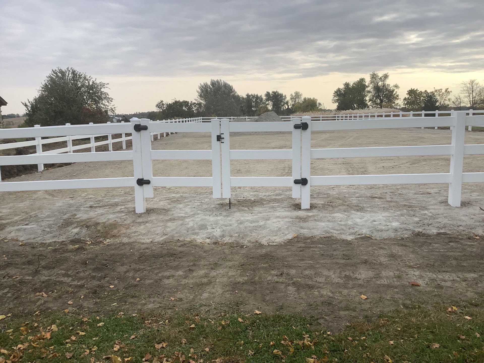 A white fence surrounds a dirt field with trees in the background