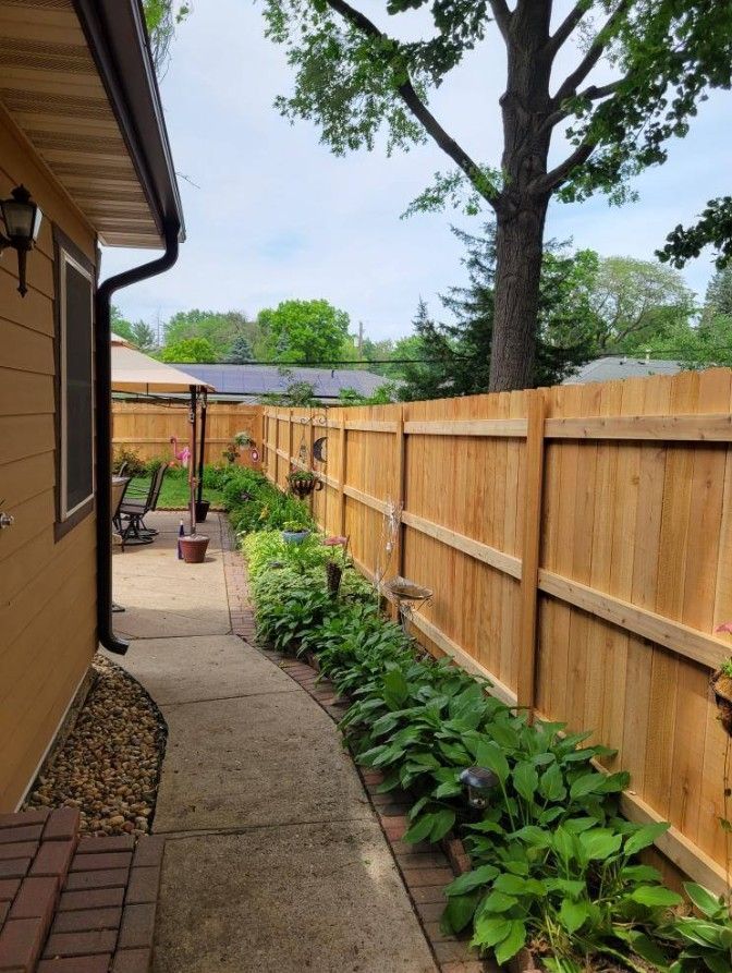 A concrete pathway curves between a house and wooden fence, lined with greenery.