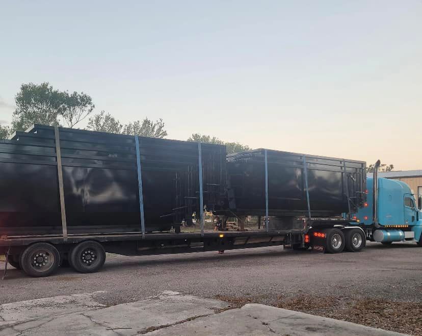 A blue semi-truck transporting two large, black cylindrical tanks on a flatbed trailer.