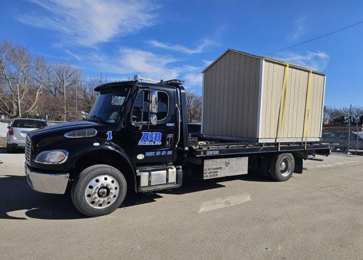 Black tow truck transporting a beige storage shed on a sunny day.