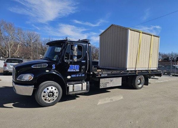 Black tow truck transporting a beige storage shed on a sunny day.