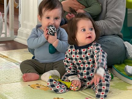 Two babies sitting on a playmat; one holds a toy and looks at it, the other looks off to the side.