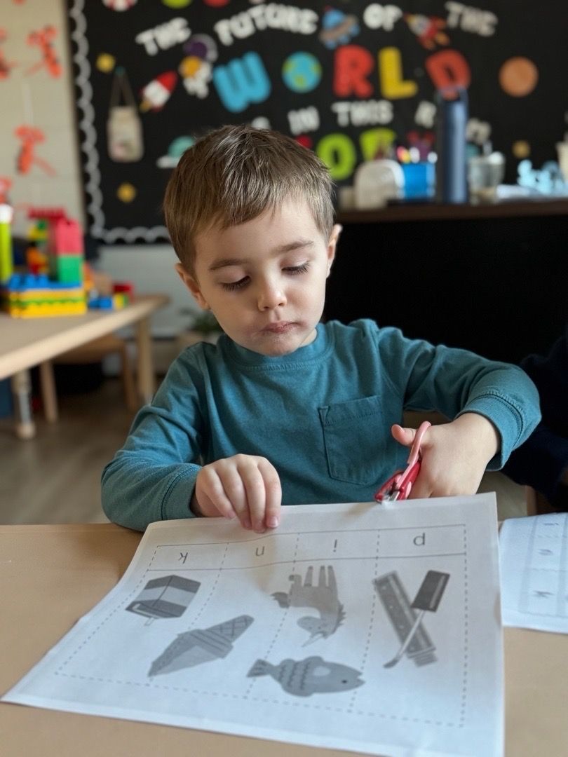 A young child sits at a table cutting shapes from a worksheet with scissors in a classroom.