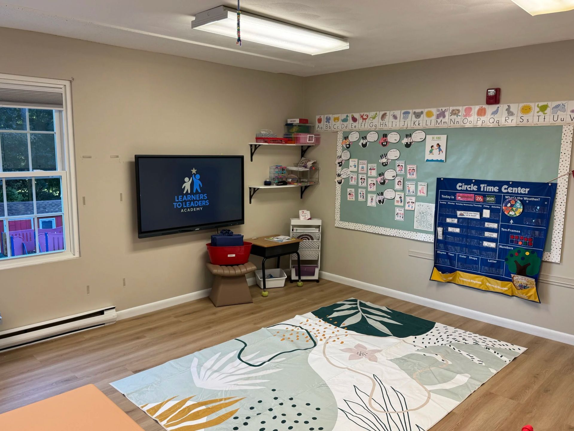 Classroom interior with TV, whiteboard, rug, and shelves. Beige walls, light wood floor, and a window.