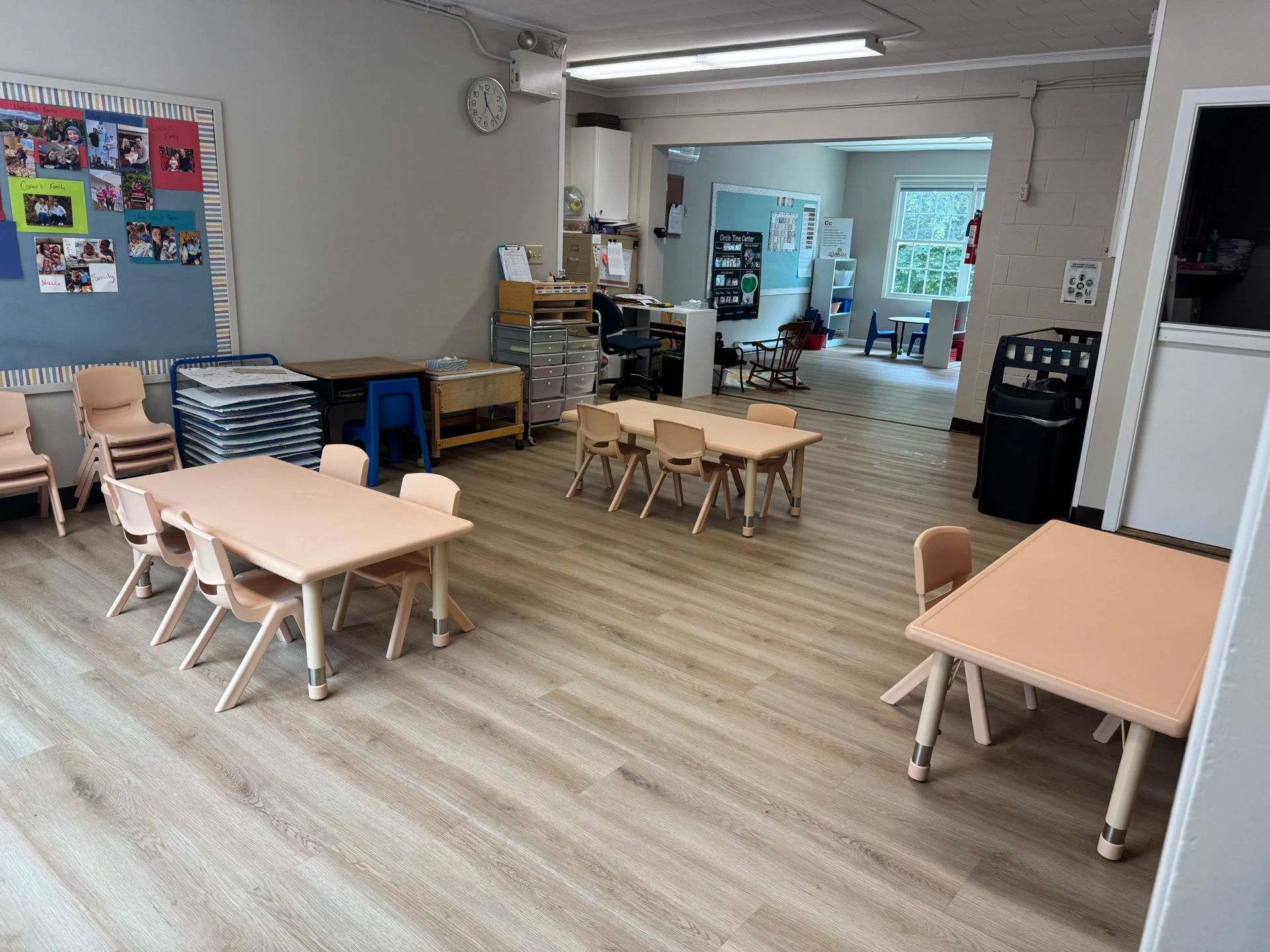 A brightly lit preschool classroom with tables and chairs, and a doorway to another area.