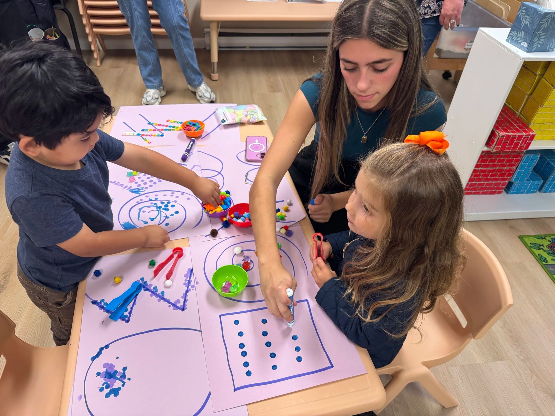 A teacher assists two children with a craft activity at a table in a classroom.