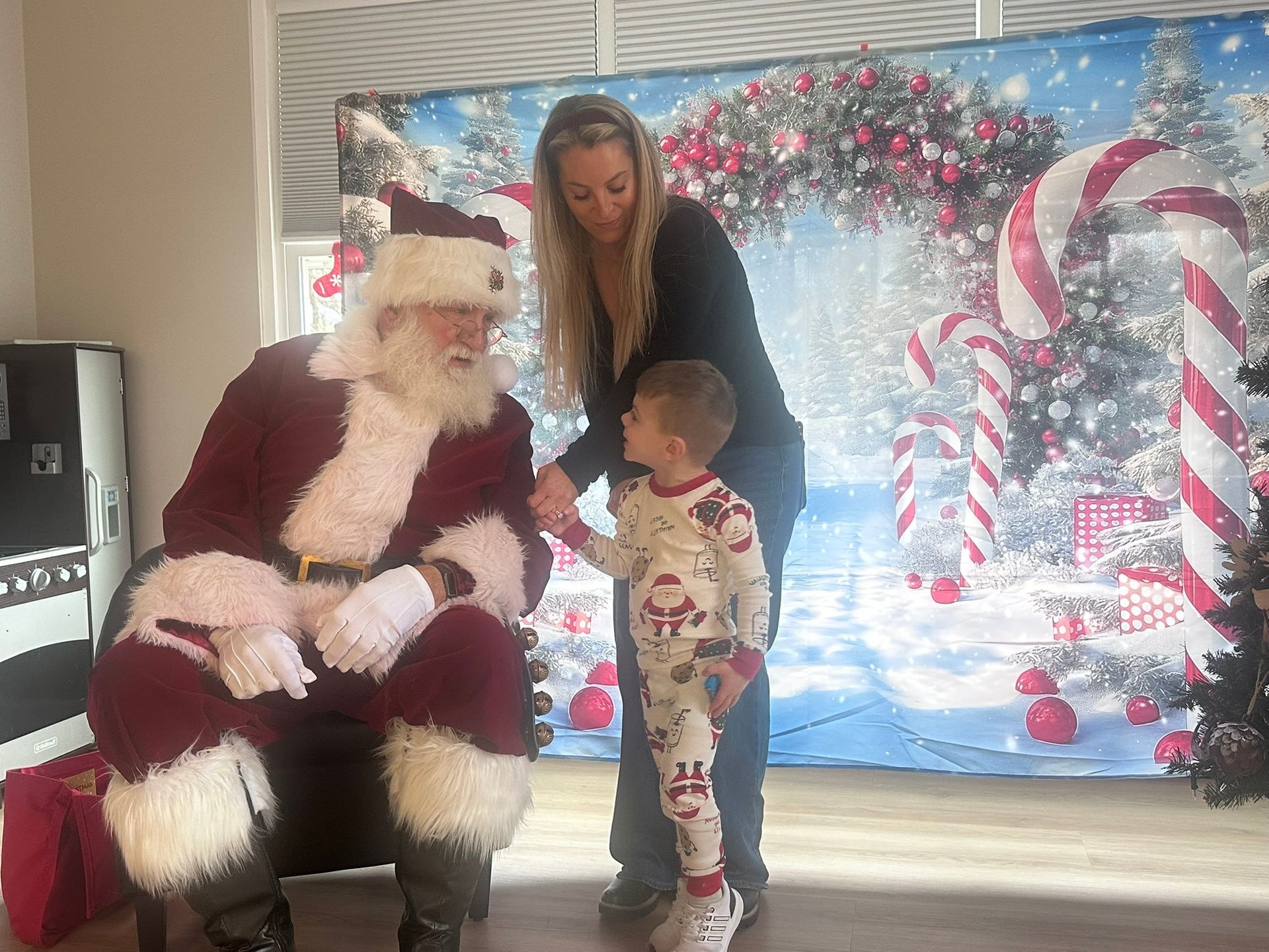 Santa Claus sitting, shaking hands with a child in pajamas, with a woman beside them, in front of a Christmas backdrop.
