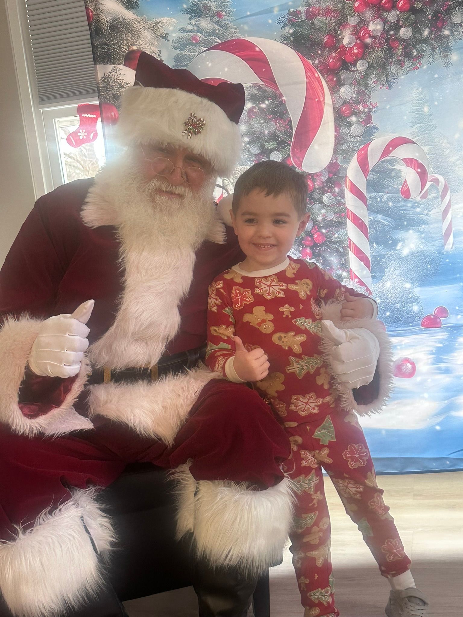 Santa Claus sits with a child wearing red pajamas, both giving thumbs up. Festive backdrop.