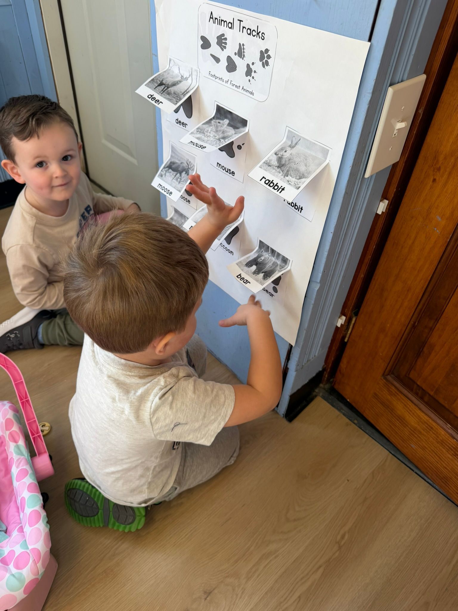 Two young children interact with a board displaying photos, one pointing, the other watching. Indoor setting.