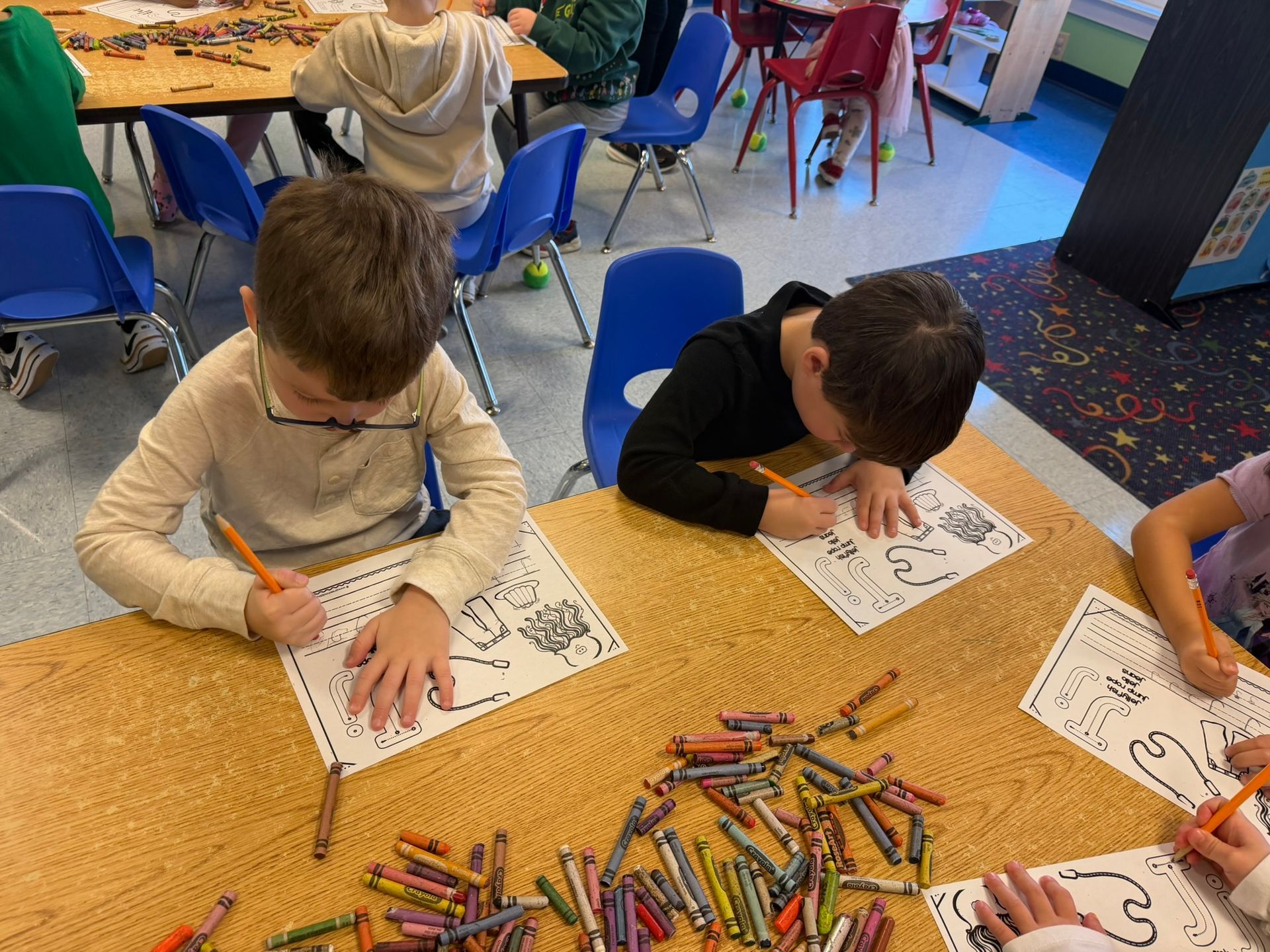 Children coloring at a table with crayons. One boy wearing glasses colors a worksheet.