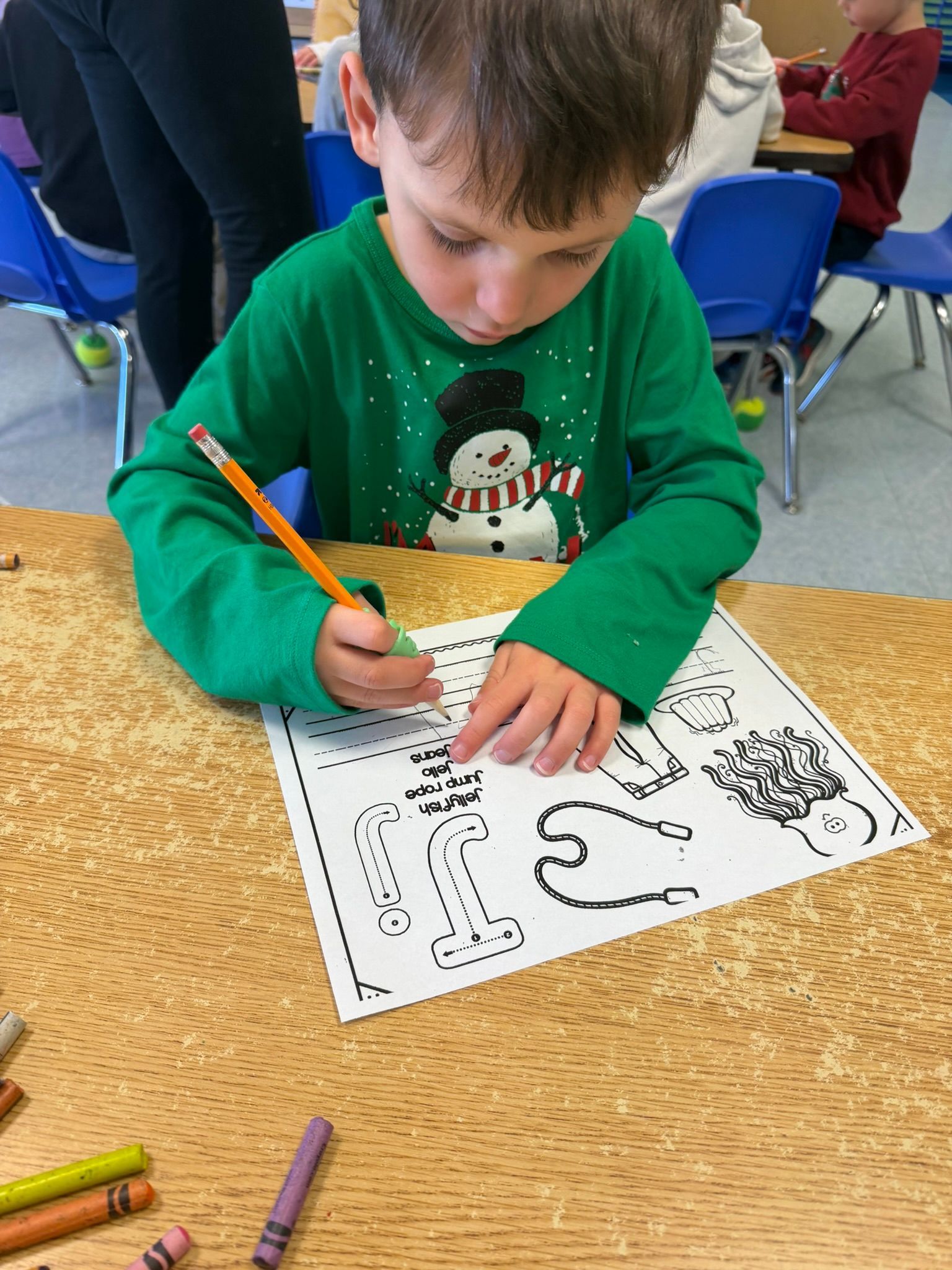 Boy coloring at a desk, wearing a green snowman sweater. Crayons are visible nearby.