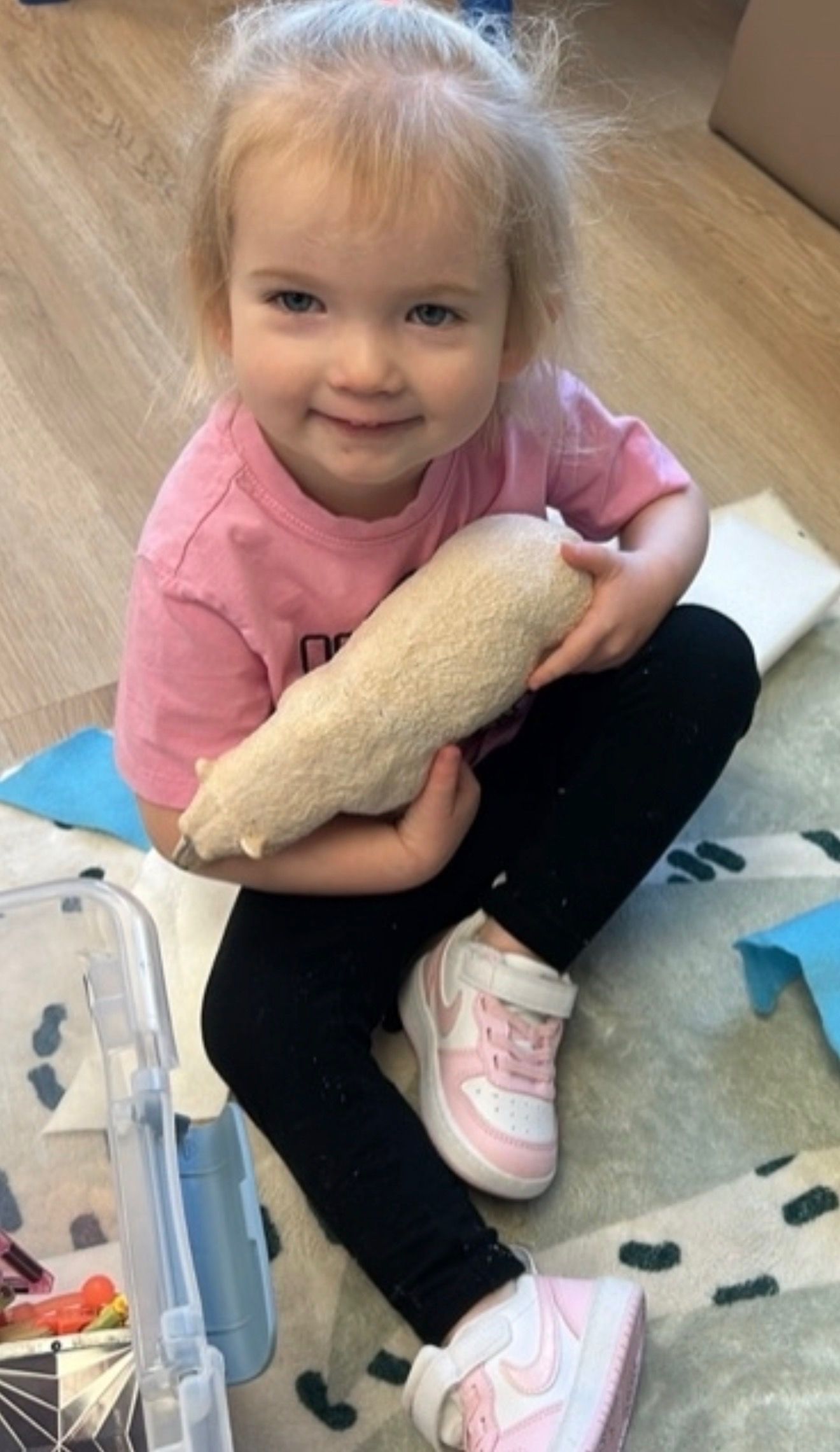 Blonde child in pink shirt and black pants, smiles, holding a beige stuffed object. Sitting on a rug.