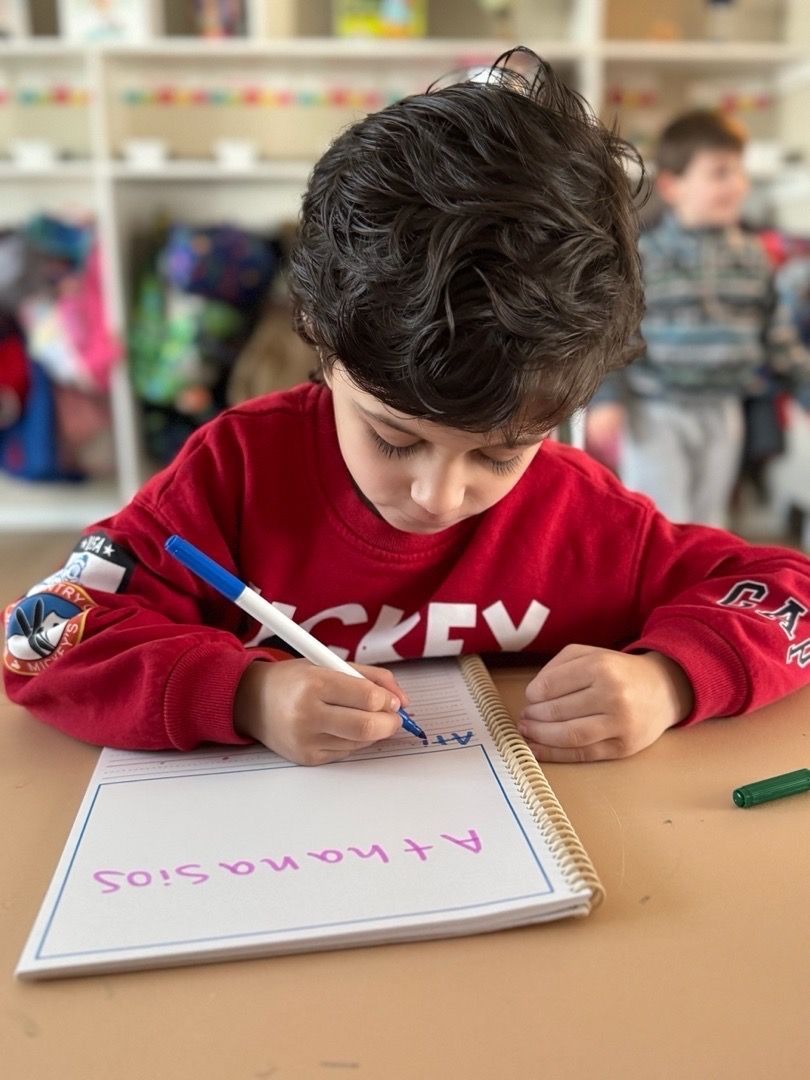Young child in red sweatshirt writing with a blue pen on a spiral notebook at a table.
