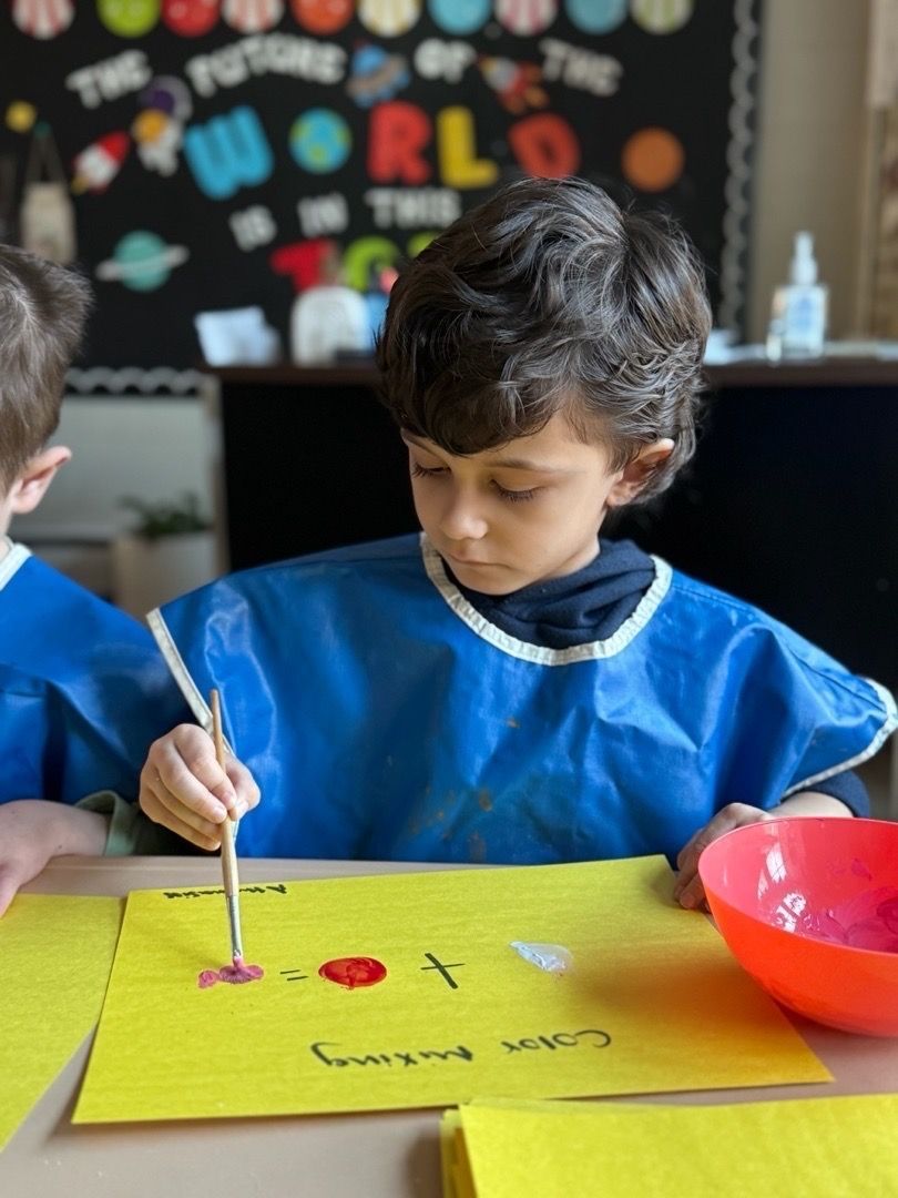 Boy painting with brush, wearing blue apron. Yellow paper, red paint, pink bowl.