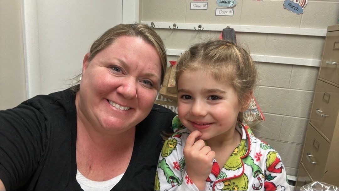 Woman and young child smiling, posing for selfie. Child wears green pajamas. Background is a classroom setting.