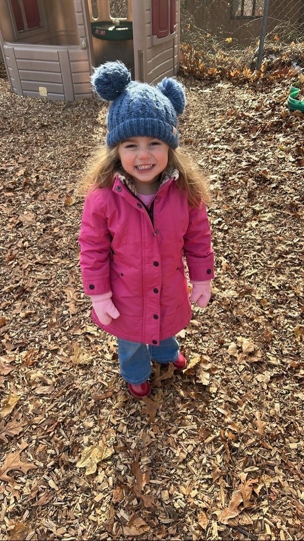Girl in pink coat and blue hat with pom poms smiles outside on a bed of leaves.