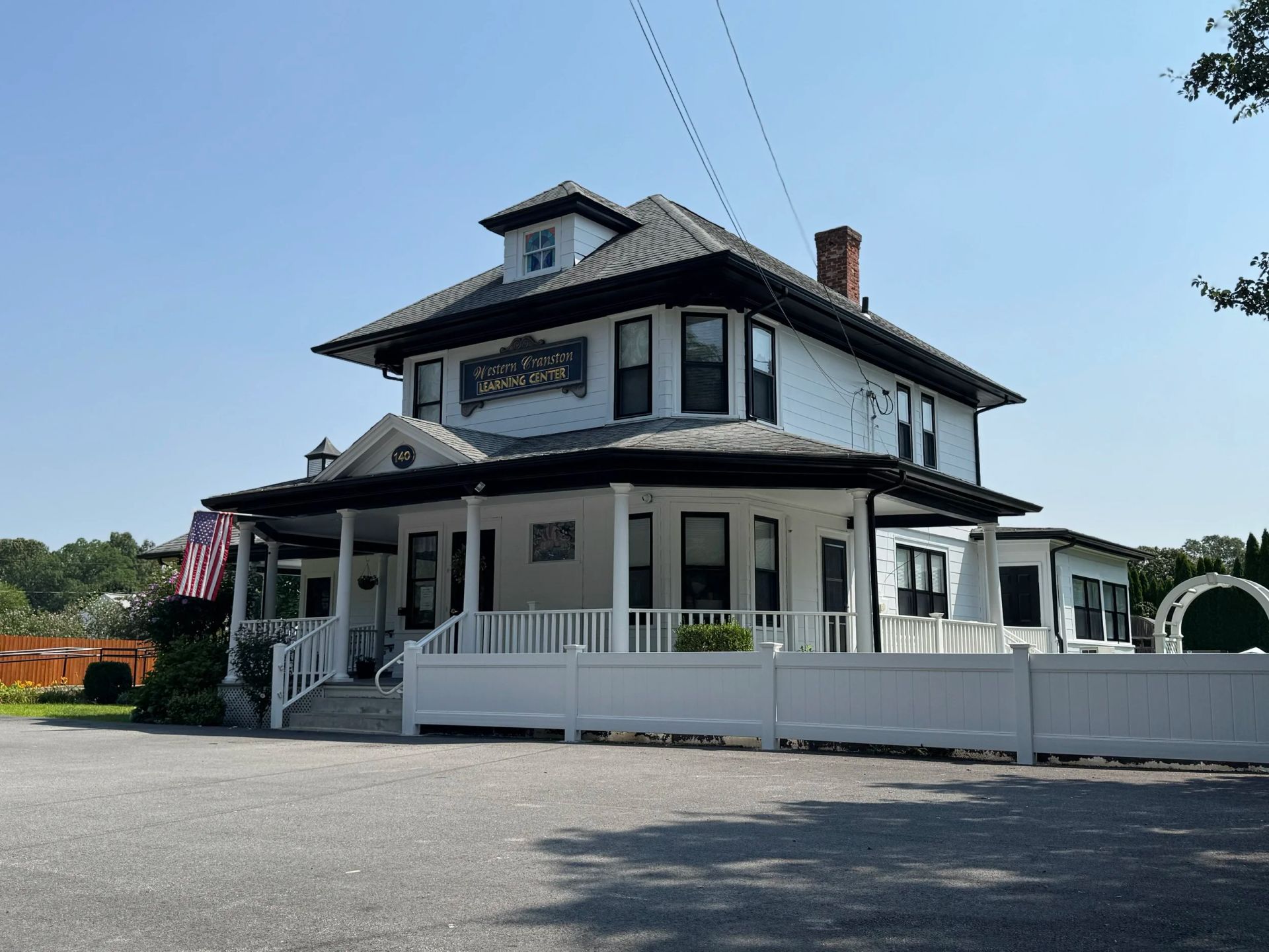 White two-story building with black trim, porch, and a fence on a sunny day.