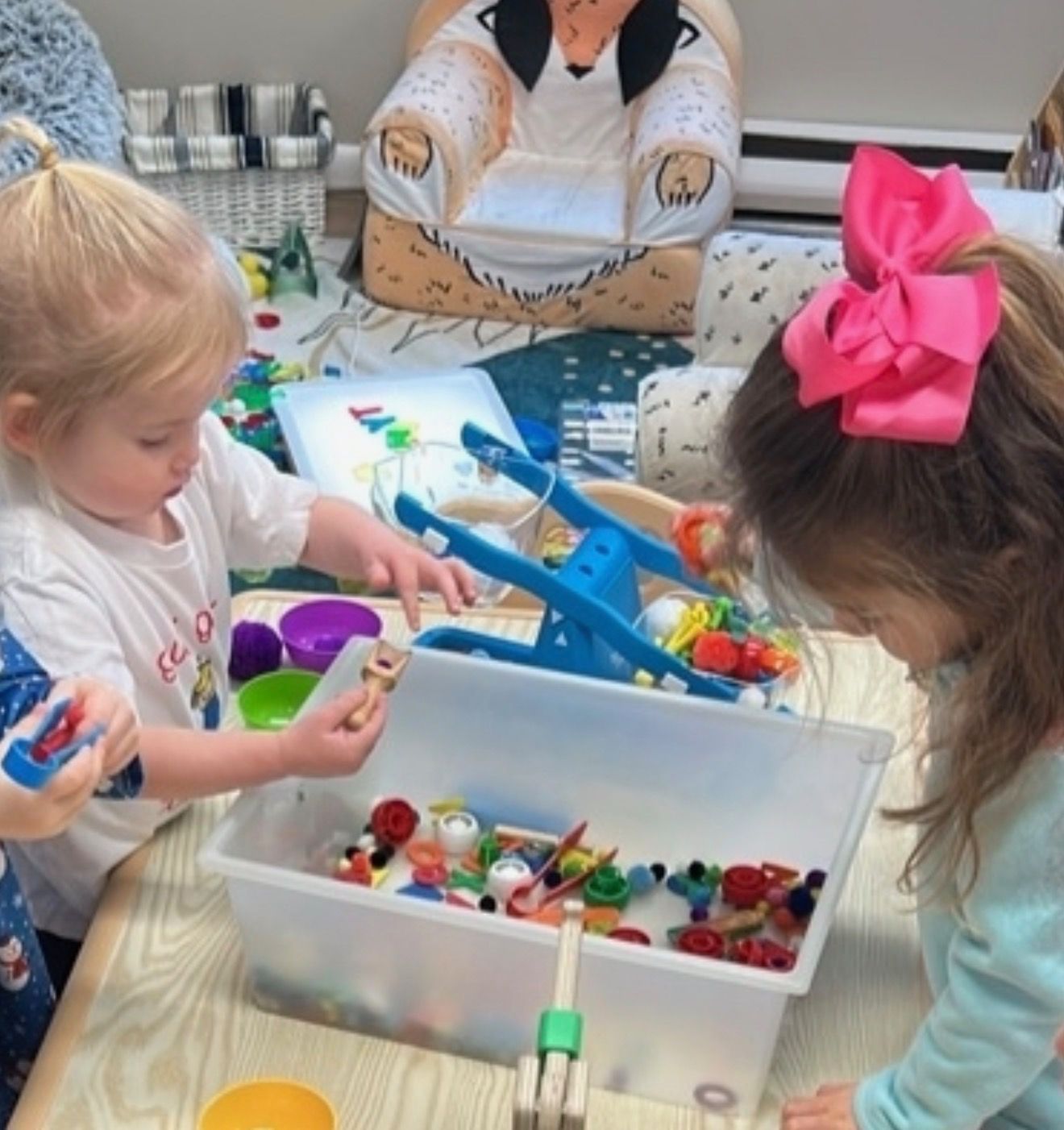 Two children playing with toys at a table; one holding a wooden peg, the other with a large pink bow.