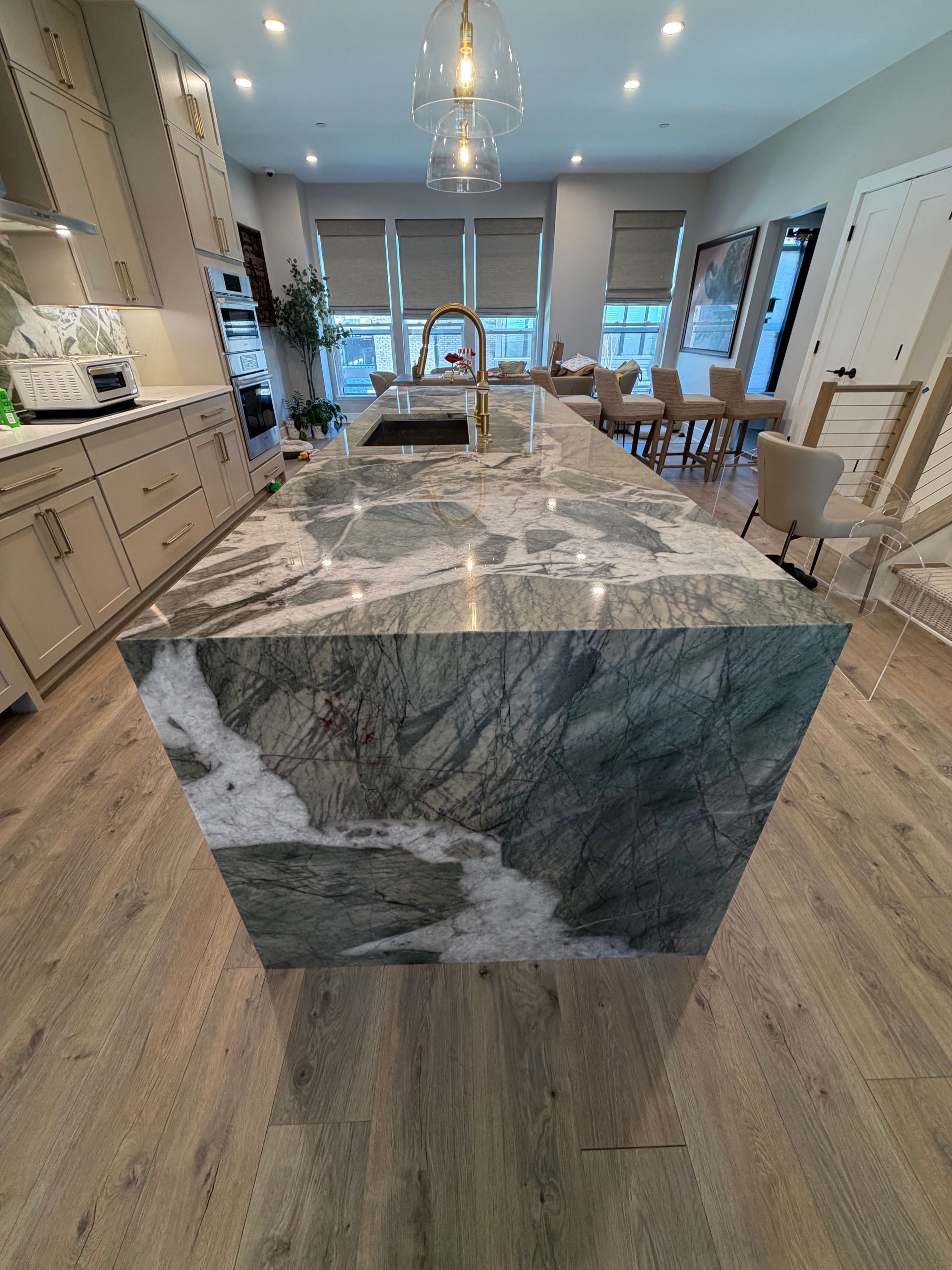 Modern kitchen with large marble island, wood flooring, and dining area near bright windows.