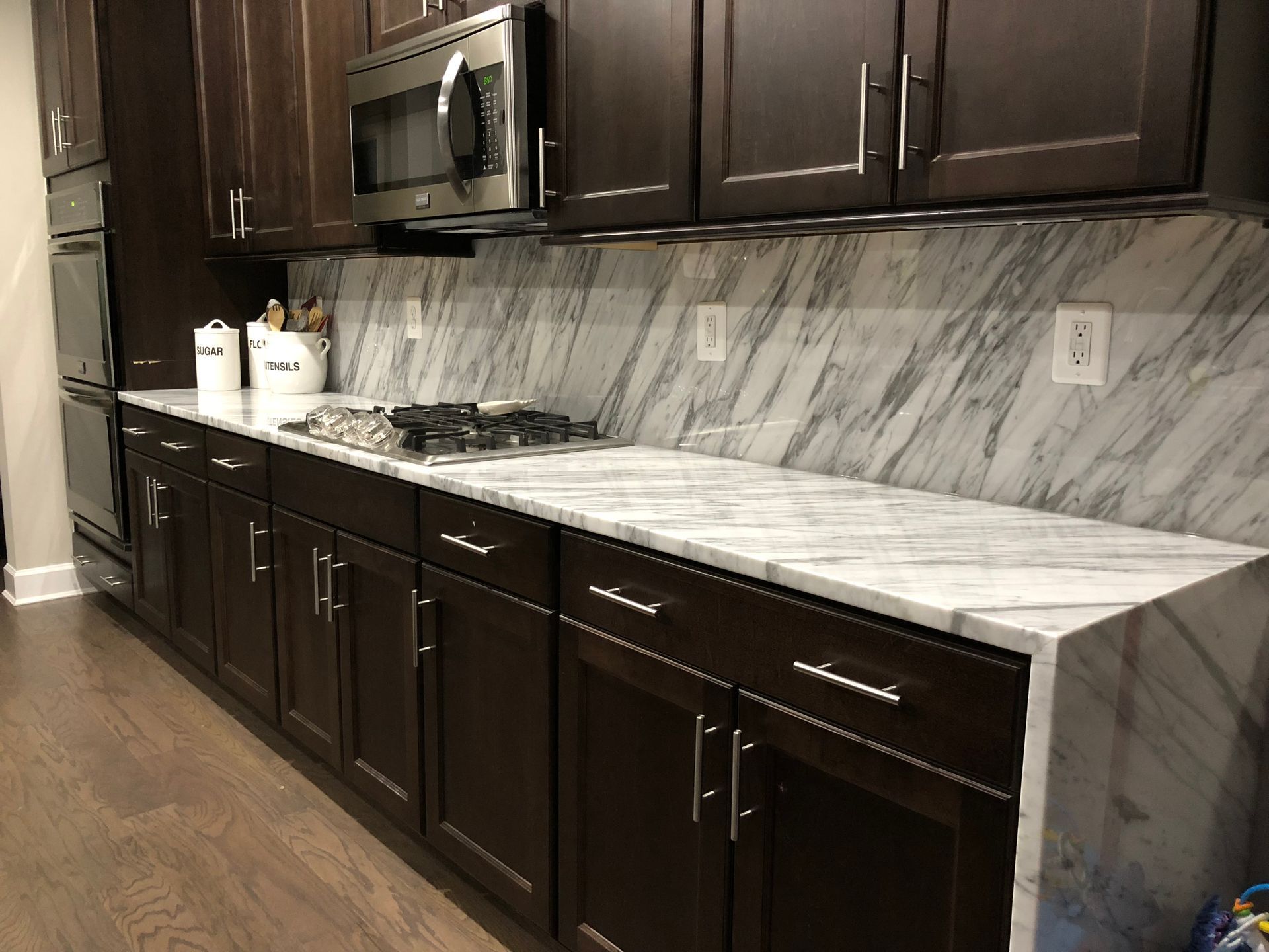 Modern kitchen with dark wood cabinets, marble countertops, stainless microwave, and herringbone tile backsplash