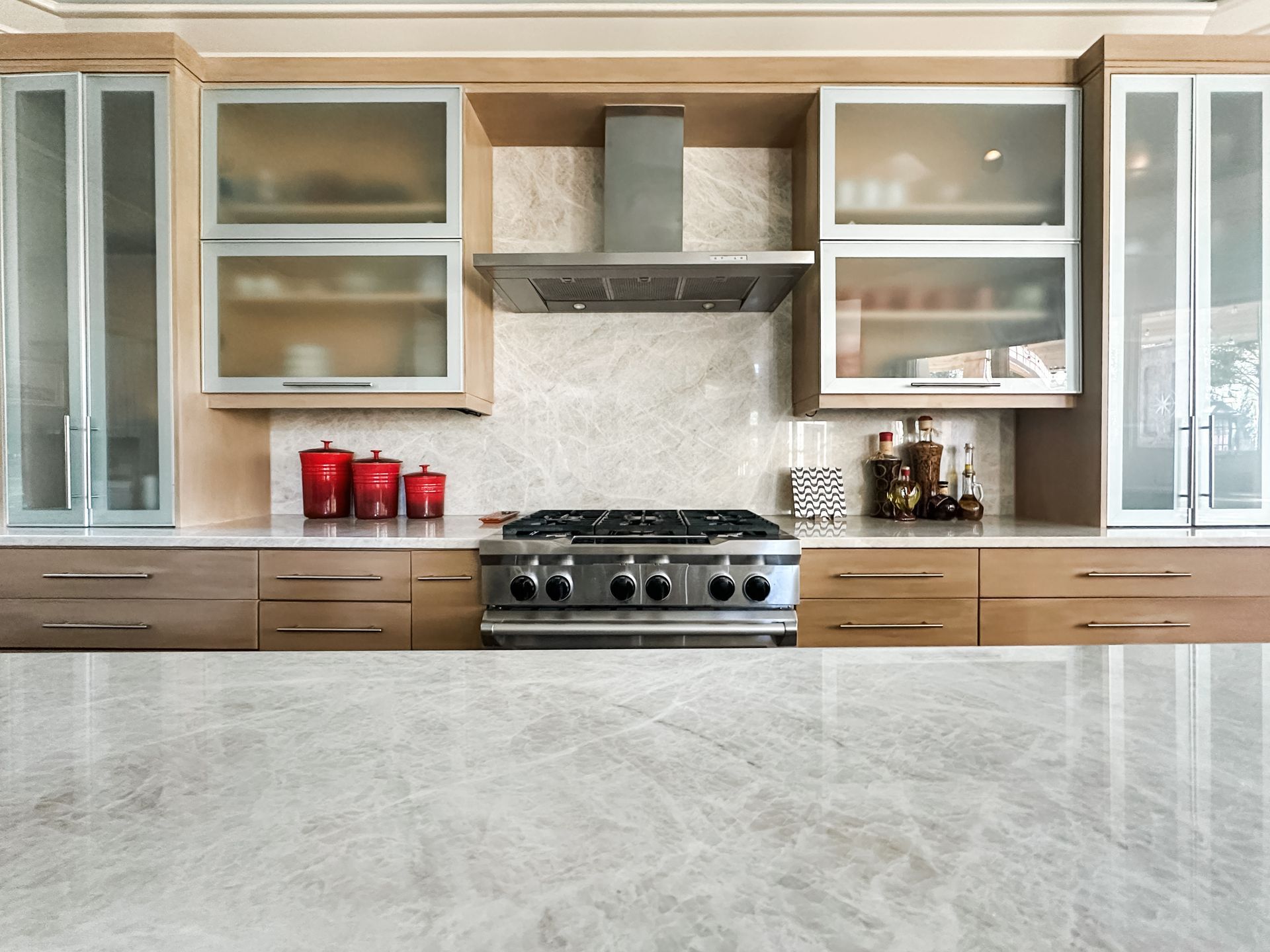 Modern kitchen with stainless steel stove, light wood cabinets, glass-front cupboards, and gray countertop