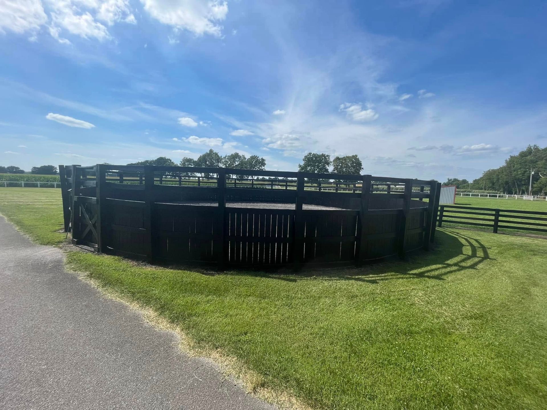Black round corral pen on grassy field beside a paved path under a blue sky
