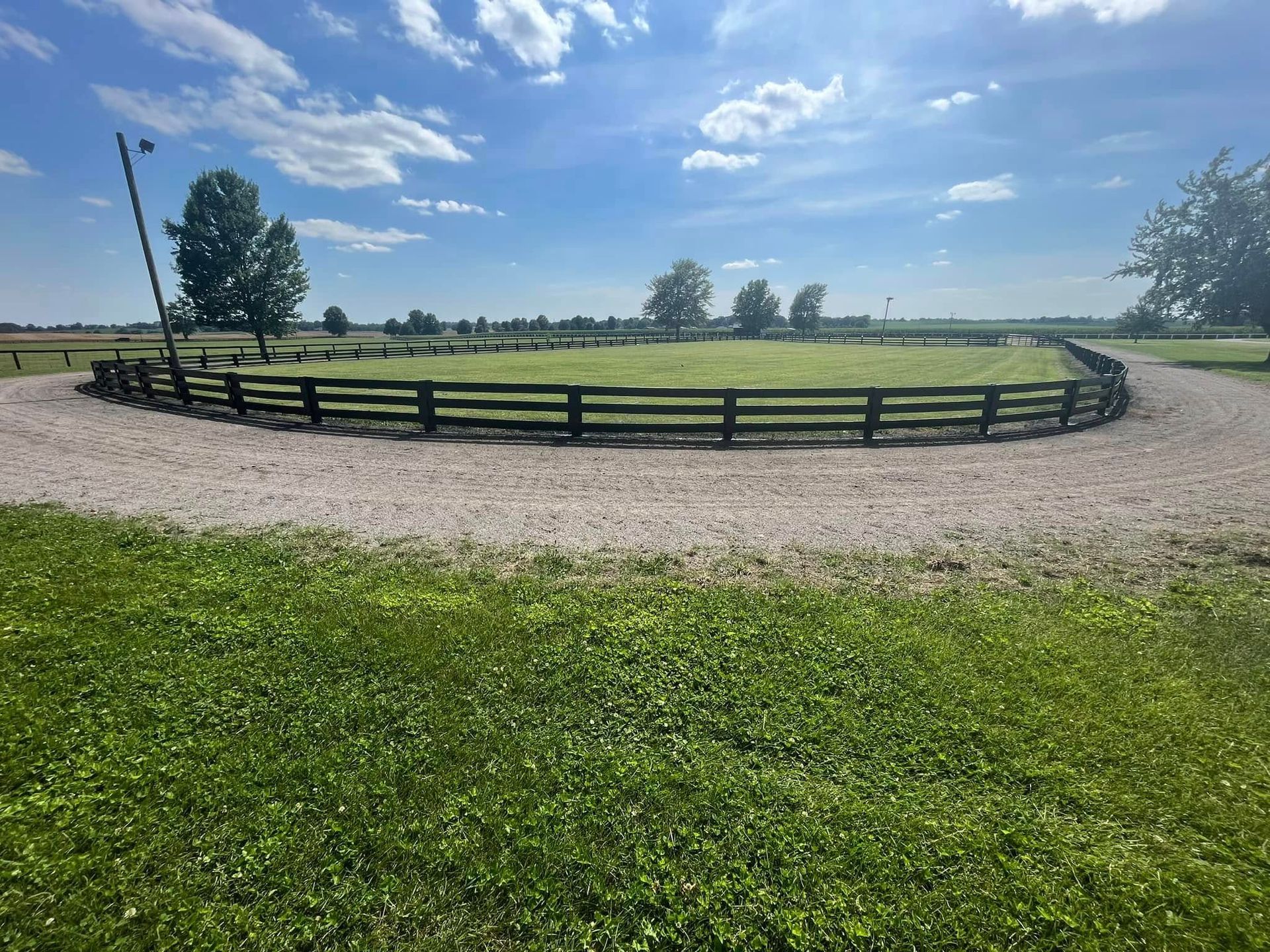 Curved wooden fence around a grassy field under a blue sky with clouds, gravel path in the foreground