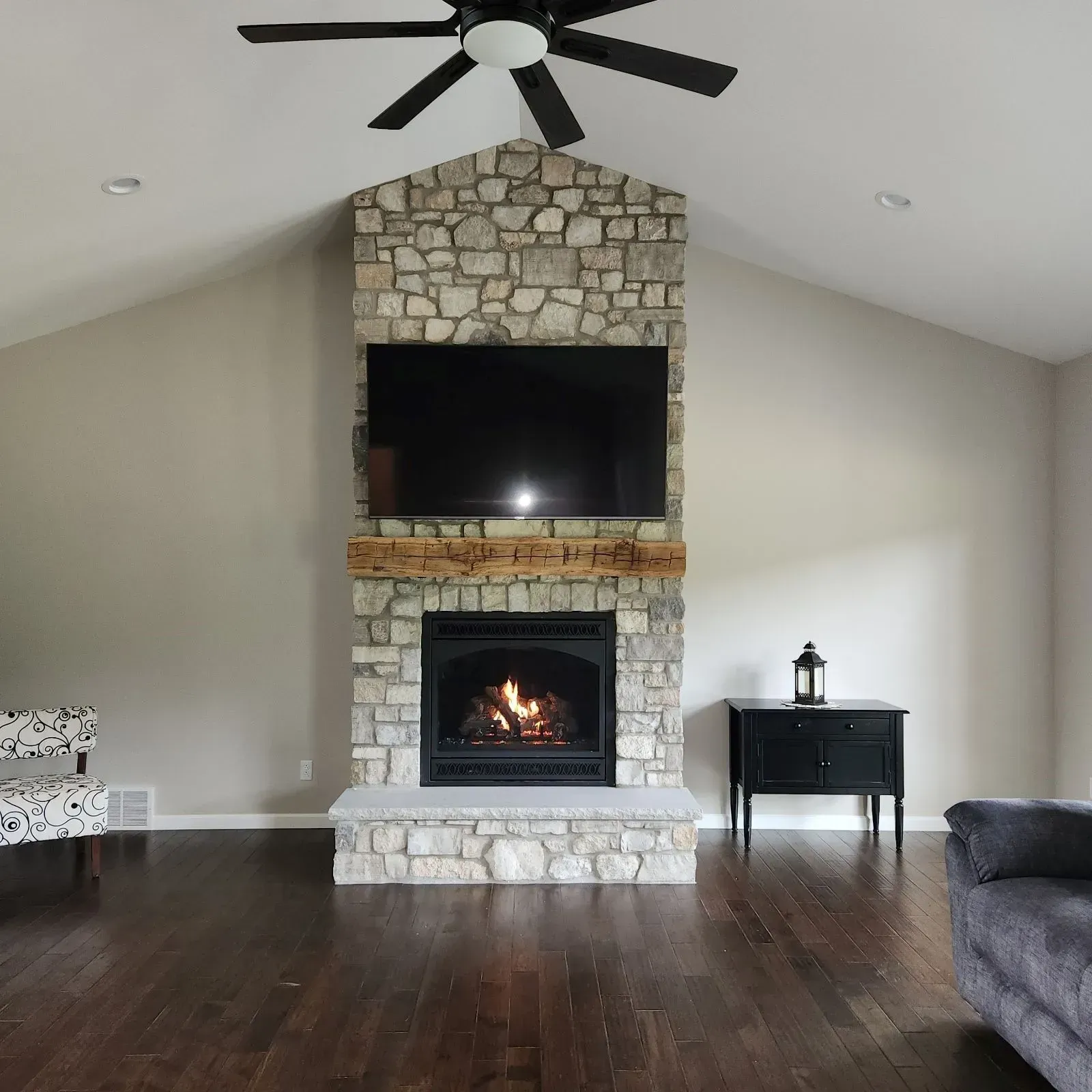 Living room with stone fireplace, TV, and black fireplace; dark wood floors and ceiling fan.