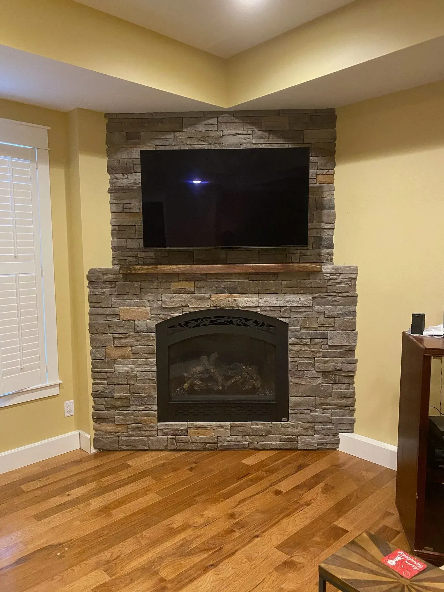 Fireplace with stone facade, TV above, in a corner of a room with wood flooring and yellow walls.