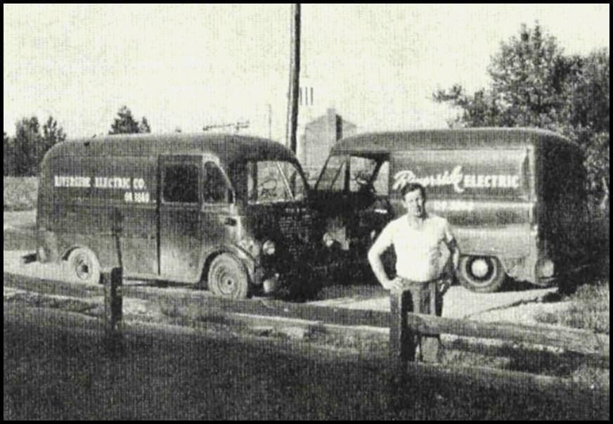 Two vintage electric delivery vans crashed roadside, with a man standing in front of them.